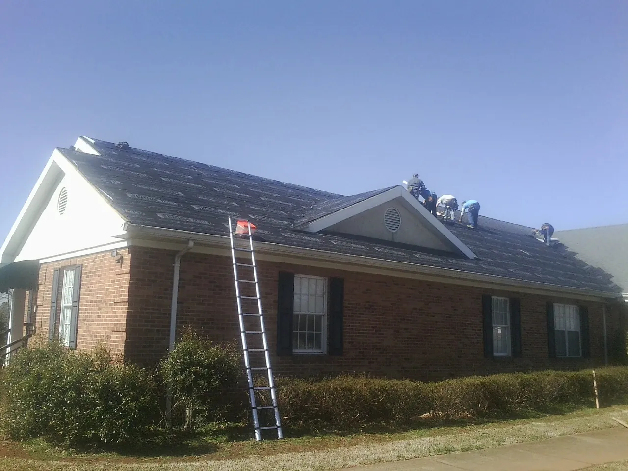 Crew tearing shingles off a brick ranch roof with a ladder set — Emergency Roof Repair work by our crew in Walnut Grove